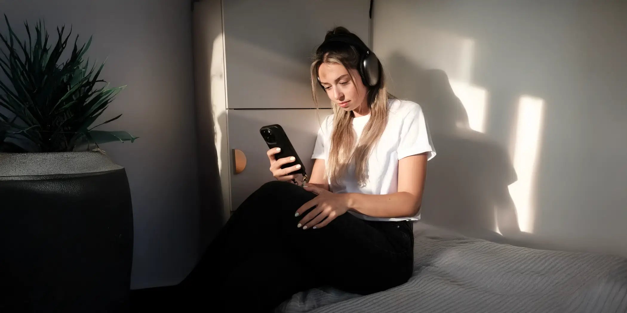 Photo d'une femme portant un casque Apple Airpods Max assise dans sa chambre et tenant un Apple iPhone 16 Pro Max muni d'une coque IONCASE dans sa main droite.