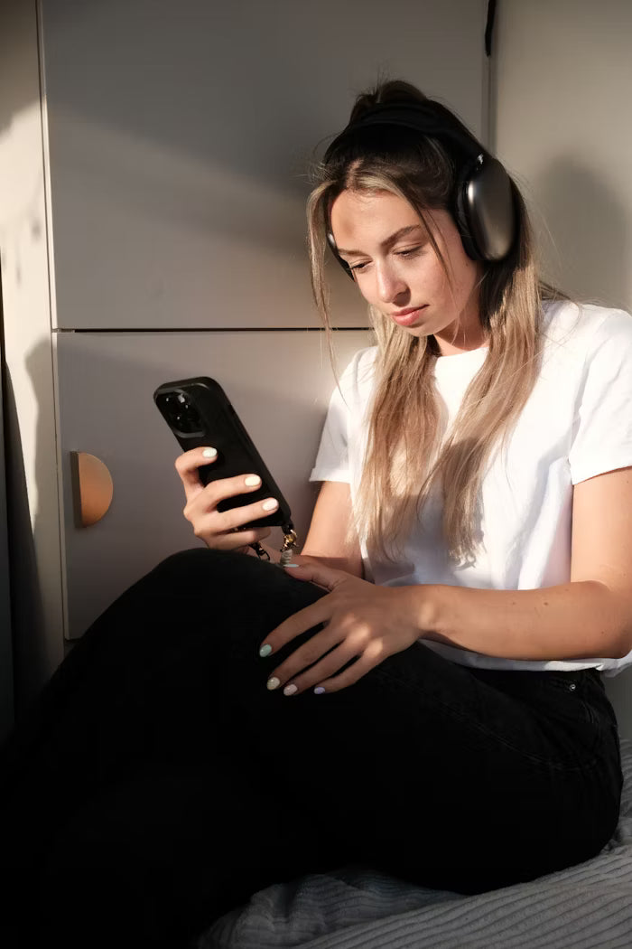 Photo d'une femme portant un casque Apple Airpods Max assise dans sa chambre et tenant un Apple iPhone 16 Pro Max muni d'une coque IONCASE dans sa main droite.